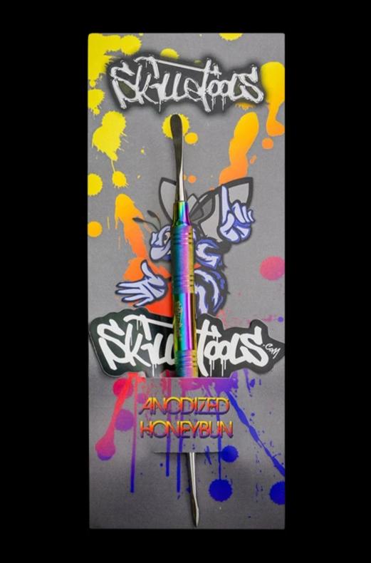 Vertical shot of a rainbow anodized Skilletools Anodized Series Dab Tools "Honeybun" on graffiti-style grey backing with yellow, orange, pink and blue paint splatters.