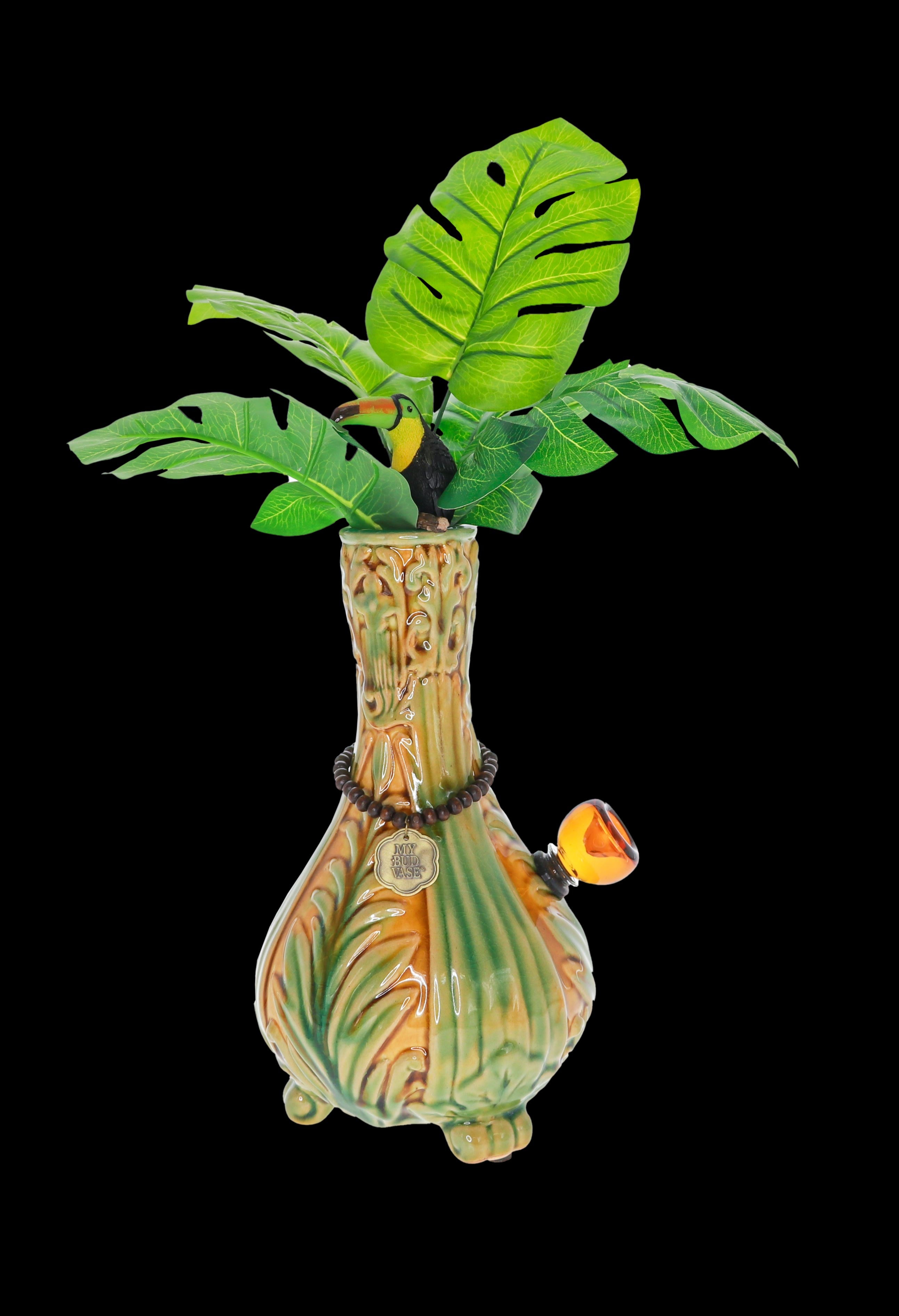 A vibrant, tropical-themed glass water pipe resembling a palm tree with a toucan perched among large green leaves, set against a black background.