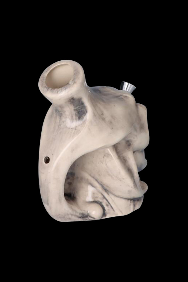 Three-quarter view of a marbled white Smiling Skull Ceramic Pipe showing its sculpted skull profile, angled mouthpiece, and metal bowl against black background.