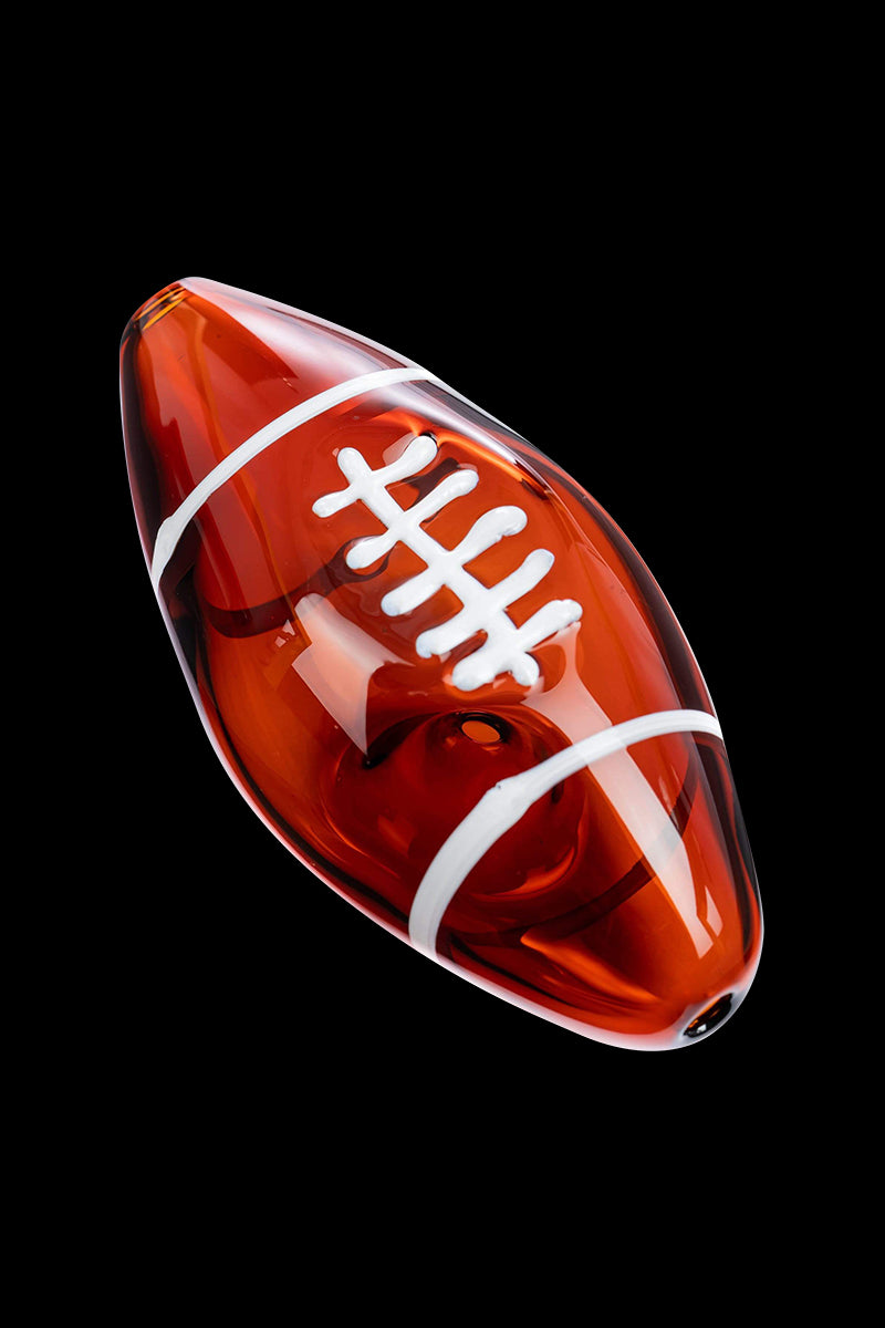 Angled close-up of the amber glass Football Hand Pipe shaped like a football with white laces and stripes against a black background