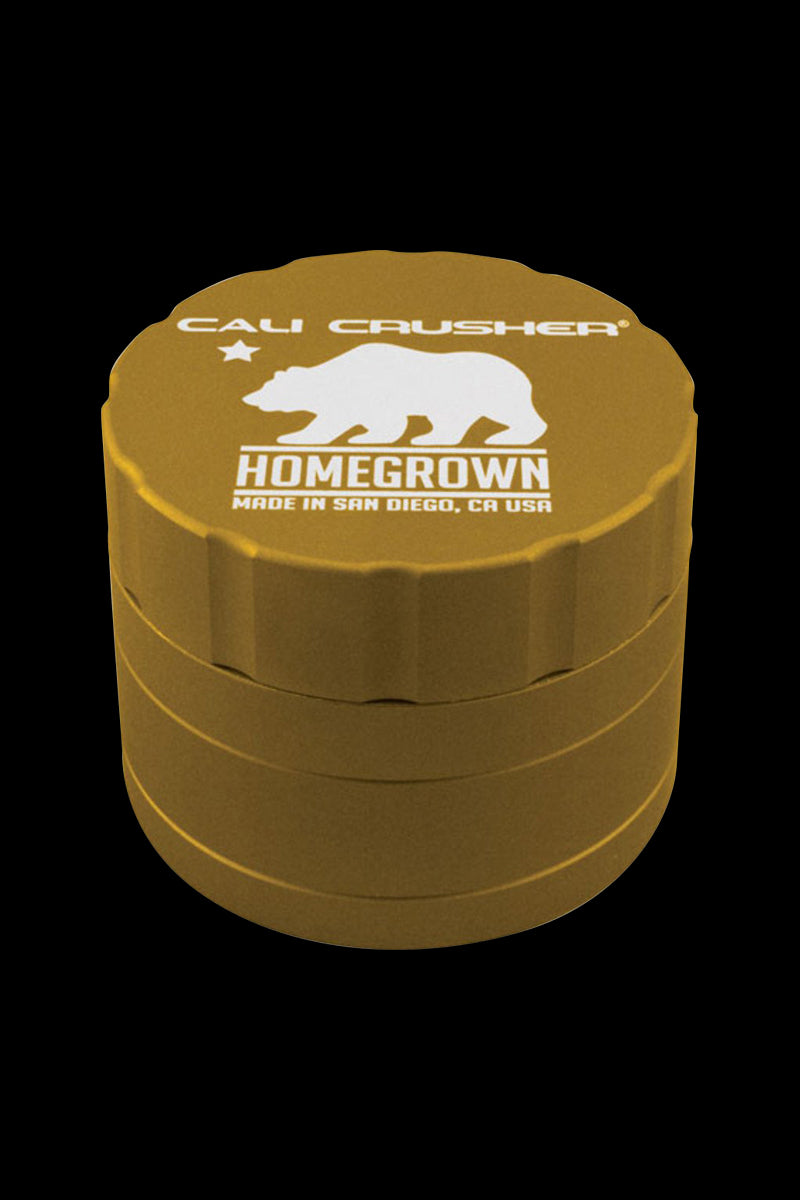 Angled close-up of the gold Cali Crusher Homegrown 4-Part Grinder showing its ridged sides and white bear logo on the lid against a black background