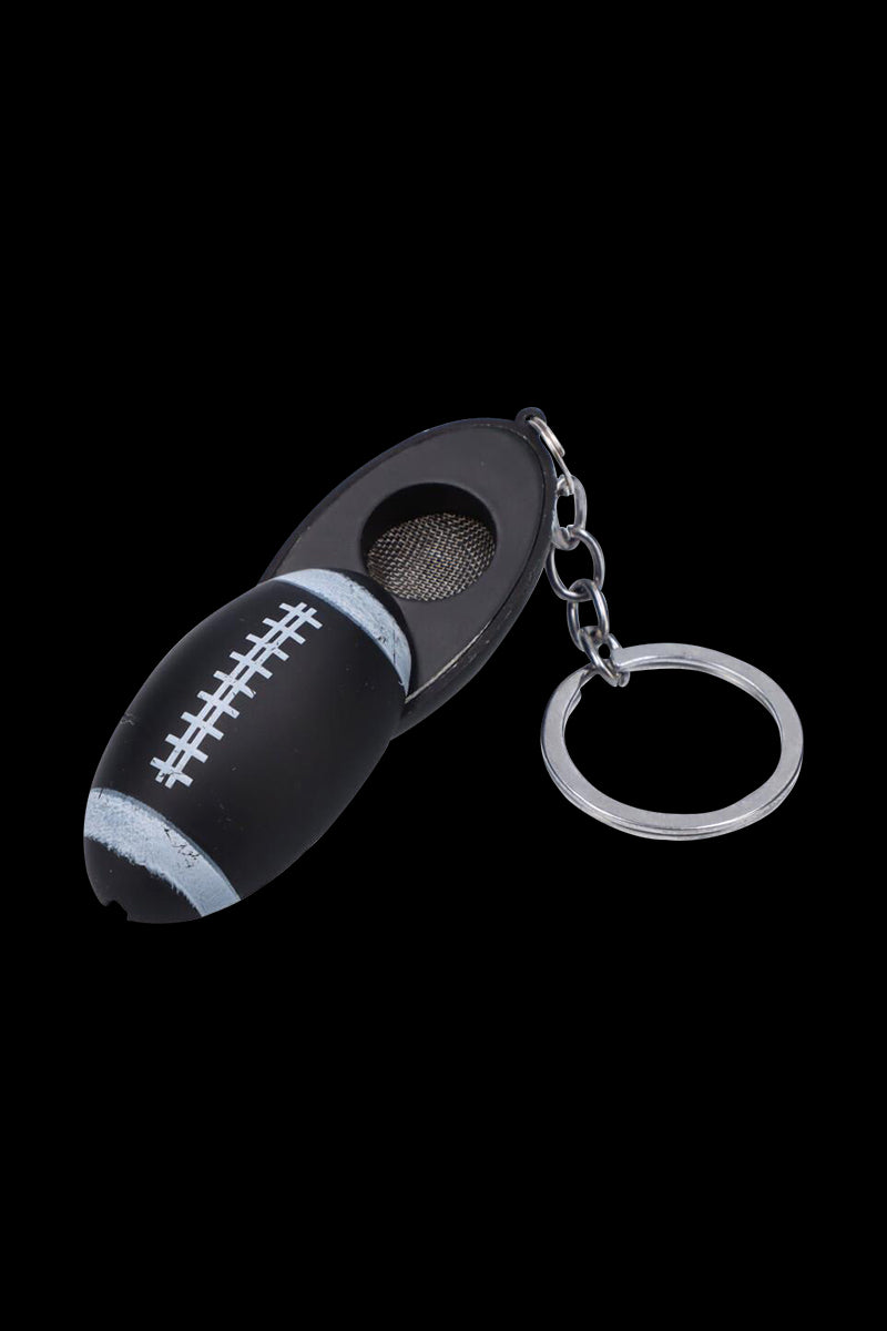 Angled close-up of the American Football Metal Hand Pipe with Key Chain showing a matte black football-shaped pipe with white laces, metal bowl, and attached silver key ring on a black background
