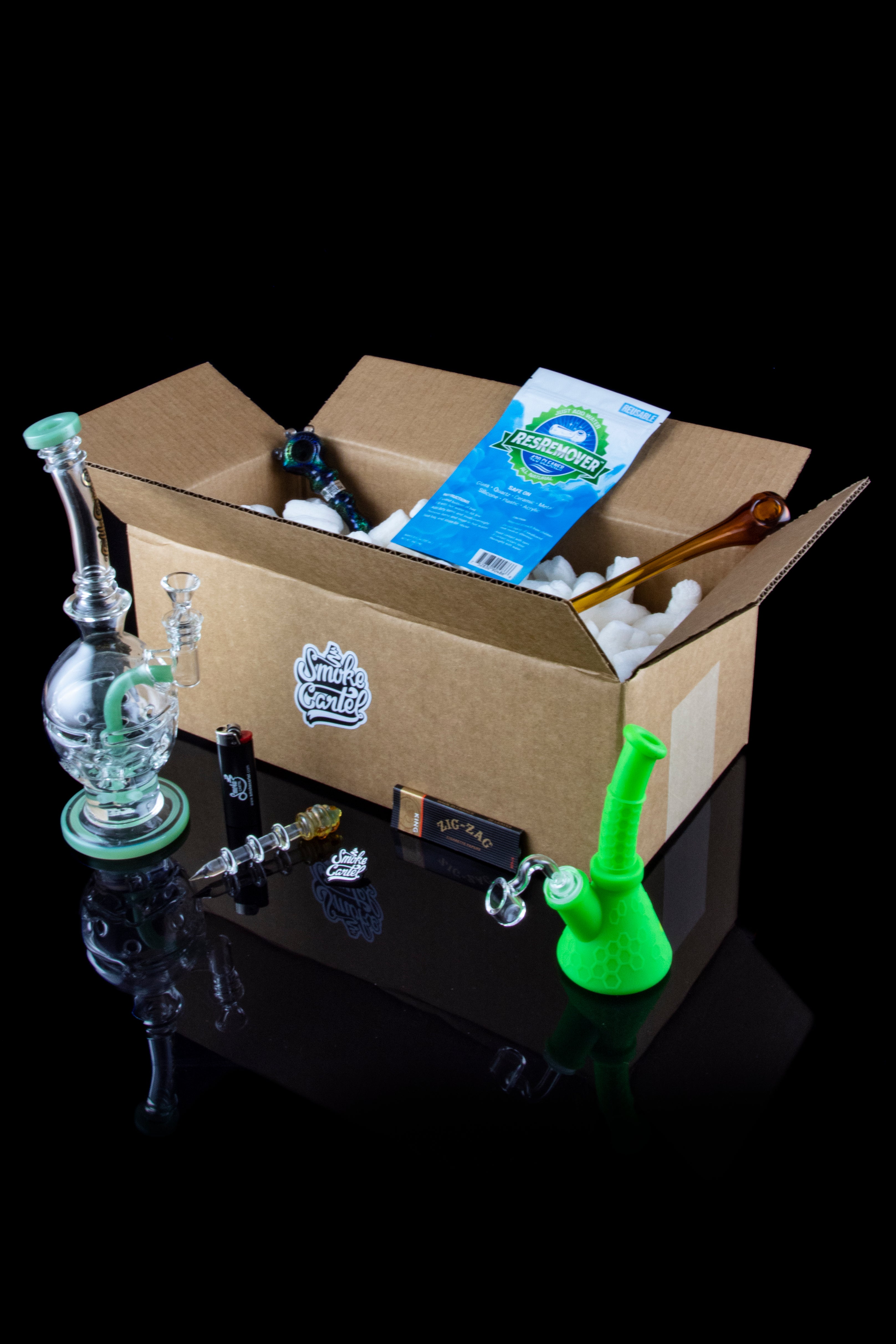 A cardboard box containing cannabis smoking accessories including a glass water pipe, a small glass bubbler, cleaning solution, and rolling papers, displayed on a reflective black surface.