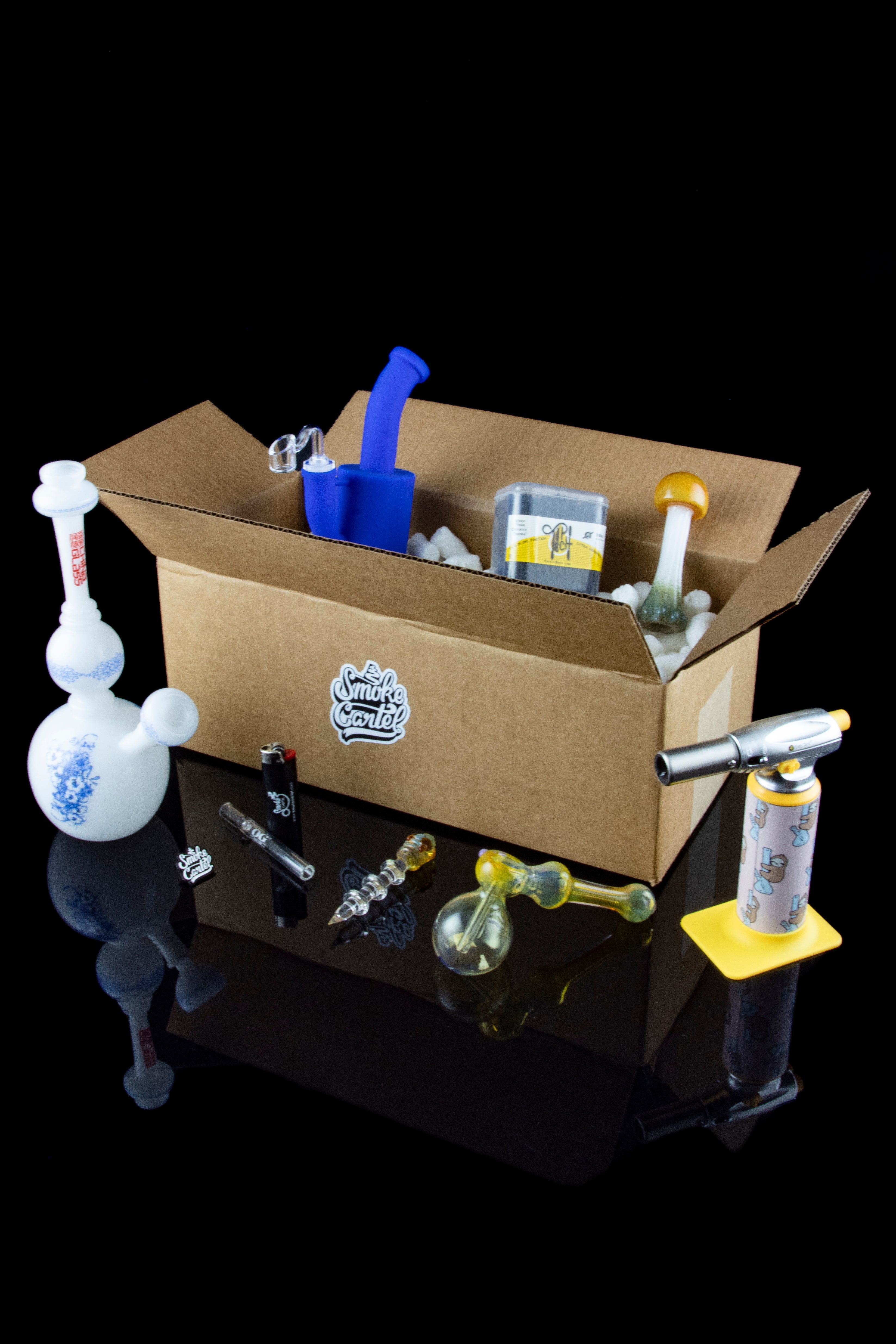 A cardboard box filled with various cannabis accessories including a water pipe (bong), dab rigs, and other smoking tools, all displayed on a reflective surface.