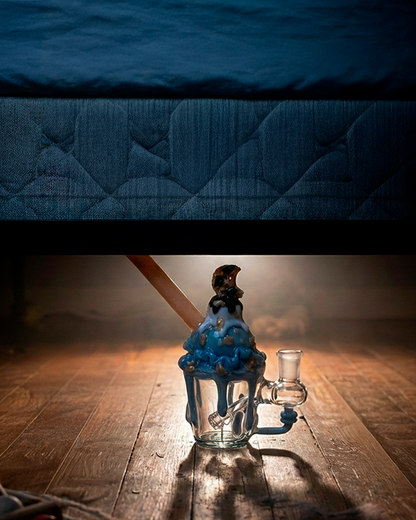 Low-angle view of the Empire Glassworks Cookie Monster Sundae Nano Rig centered on a worn wooden floor, blue glaze drips and a scoop-shaped mouthpiece lit from behind