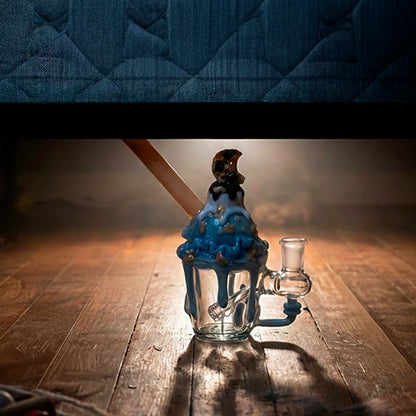 Low-angle shot of the Empire Glassworks Cookie Monster Sundae Nano Rig on a wooden floor with warm backlighting, the blue "sundae" drips and cookie topper clearly visible.