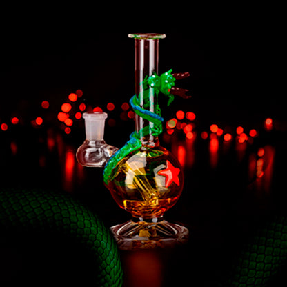 Center-front view of the Empire Glassworks Dragon Sphere Mini-Tube with a green glass dragon spiraling around a glowing amber globe against red bokeh lights