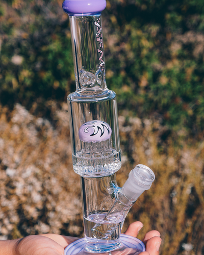 A clear glass water pipe (bong) with a purple mouthpiece and base, held in a hand against a blurred natural background.