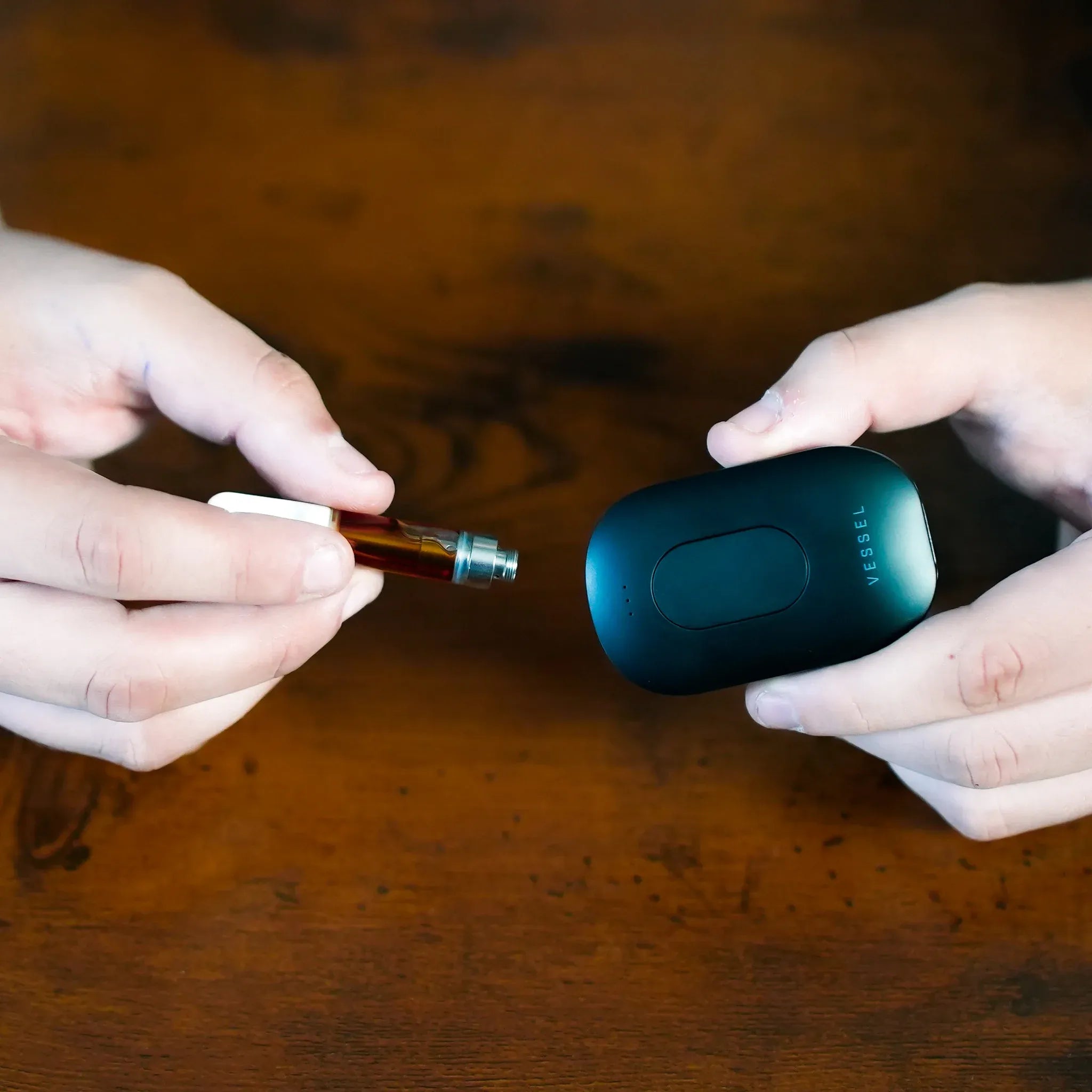 Close-up of hands connecting a cartridge to the teal Freeze Pipe Vape Pen against a warm wood tabletop.