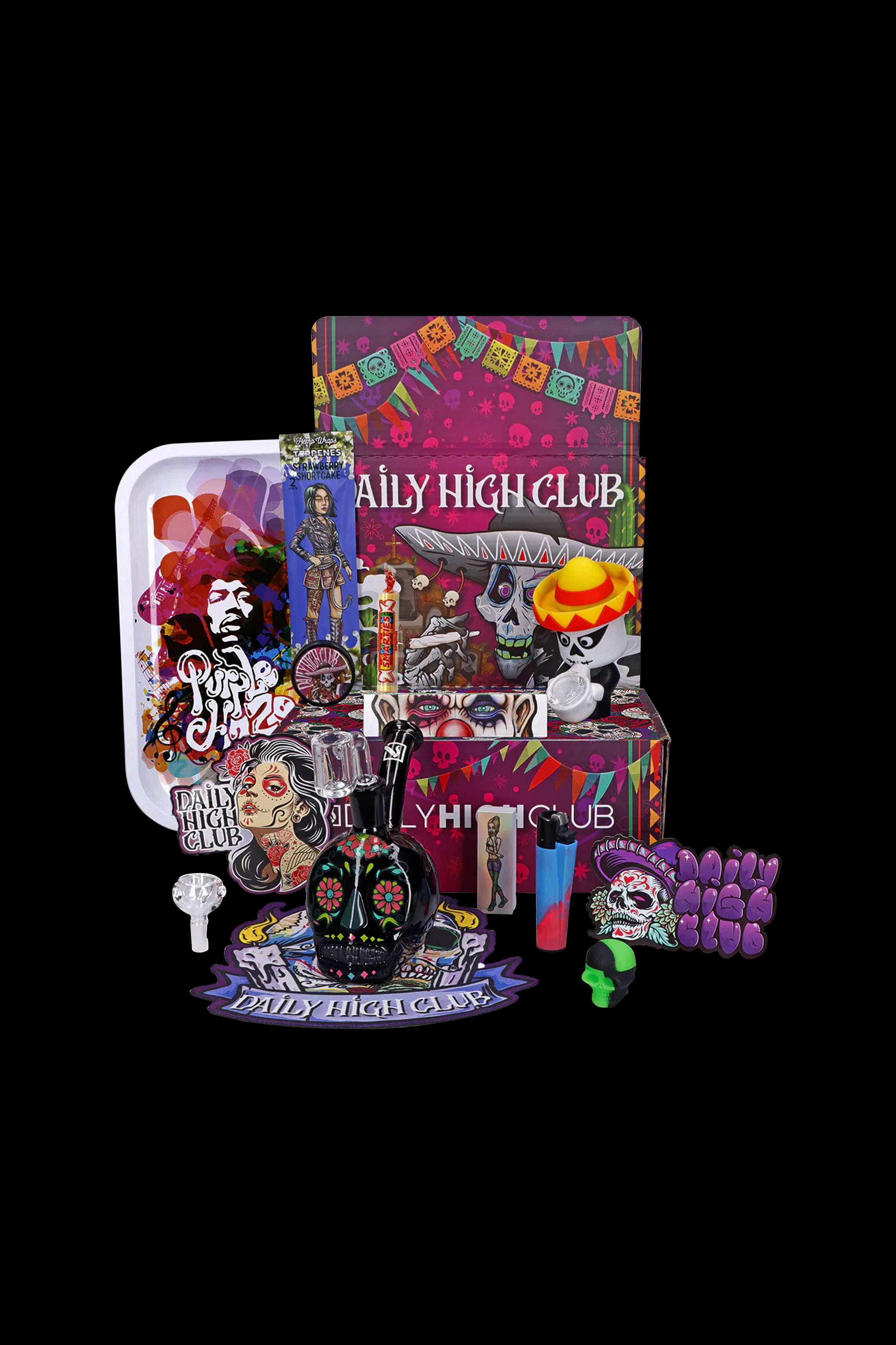 Day of the Dead themed cannabis subscription box with rolling papers, grinder, smoking accessories, and colorful glass pieces displayed on a white surface against a dark background.