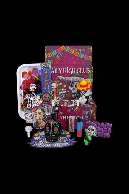 Day of the Dead themed cannabis subscription box with rolling papers, grinder, smoking accessories, and colorful glass pieces displayed on a white surface against a dark background.
