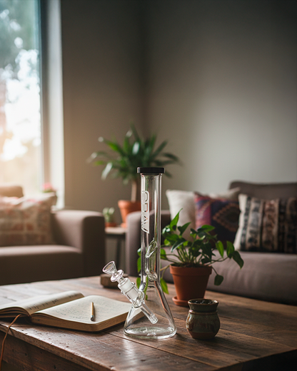 Straight-on view of the GRAV¬Æ Medium Black Accent Beaker Base Bong centered on a wooden coffee table with a plant, open notebook, and cozy living room background.