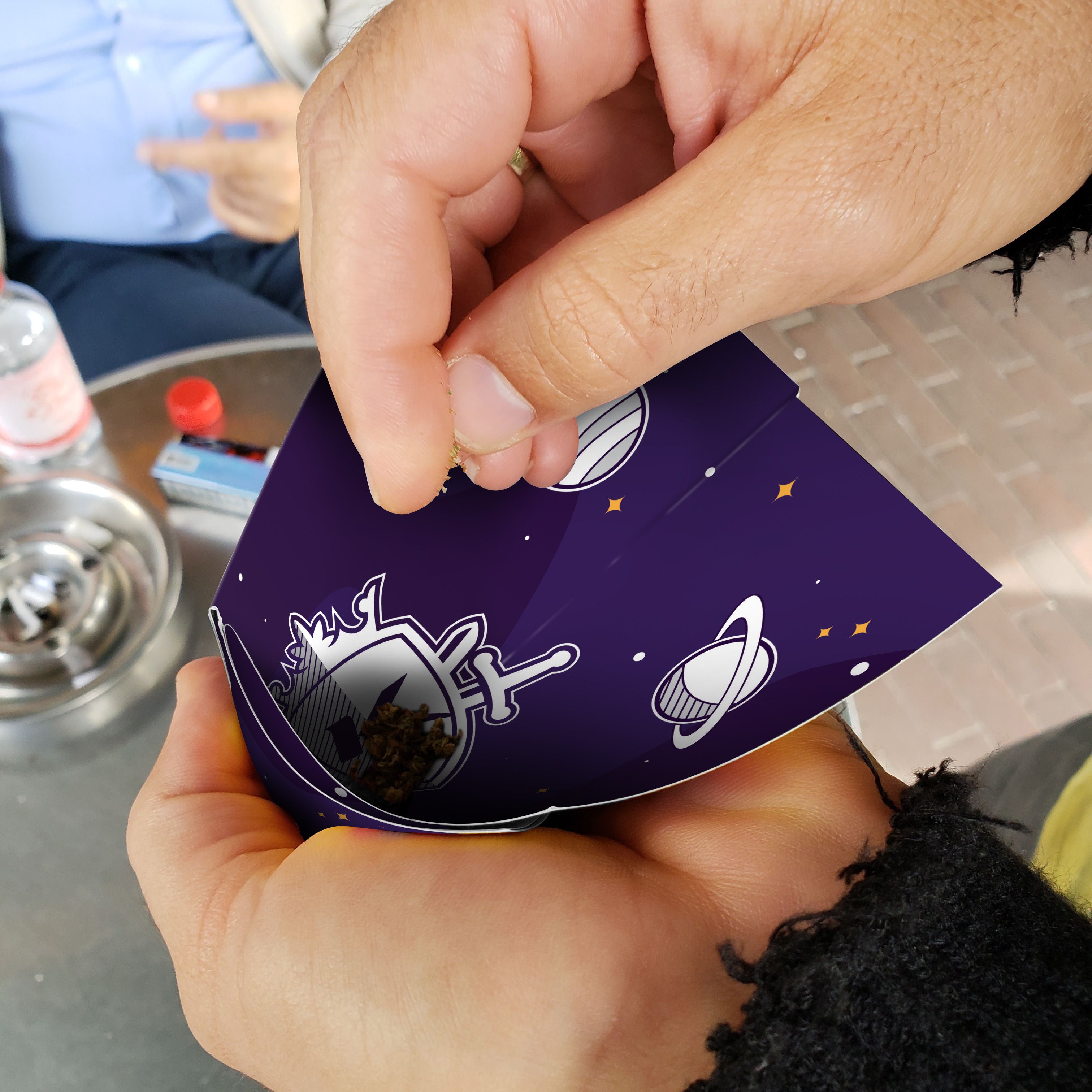 Hands holding a patterned cannabis-themed card with space imagery, near a grinder and smoking accessories.
