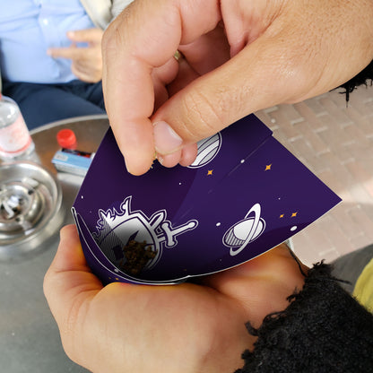 Hands holding a patterned cannabis-themed card with space imagery, near a grinder and smoking accessories.
