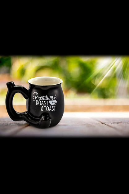 White ceramic mug with “Premium Roast & Toast” lettering and a cannabis leaf graphic, displayed on a wood surface with a blurred green background.