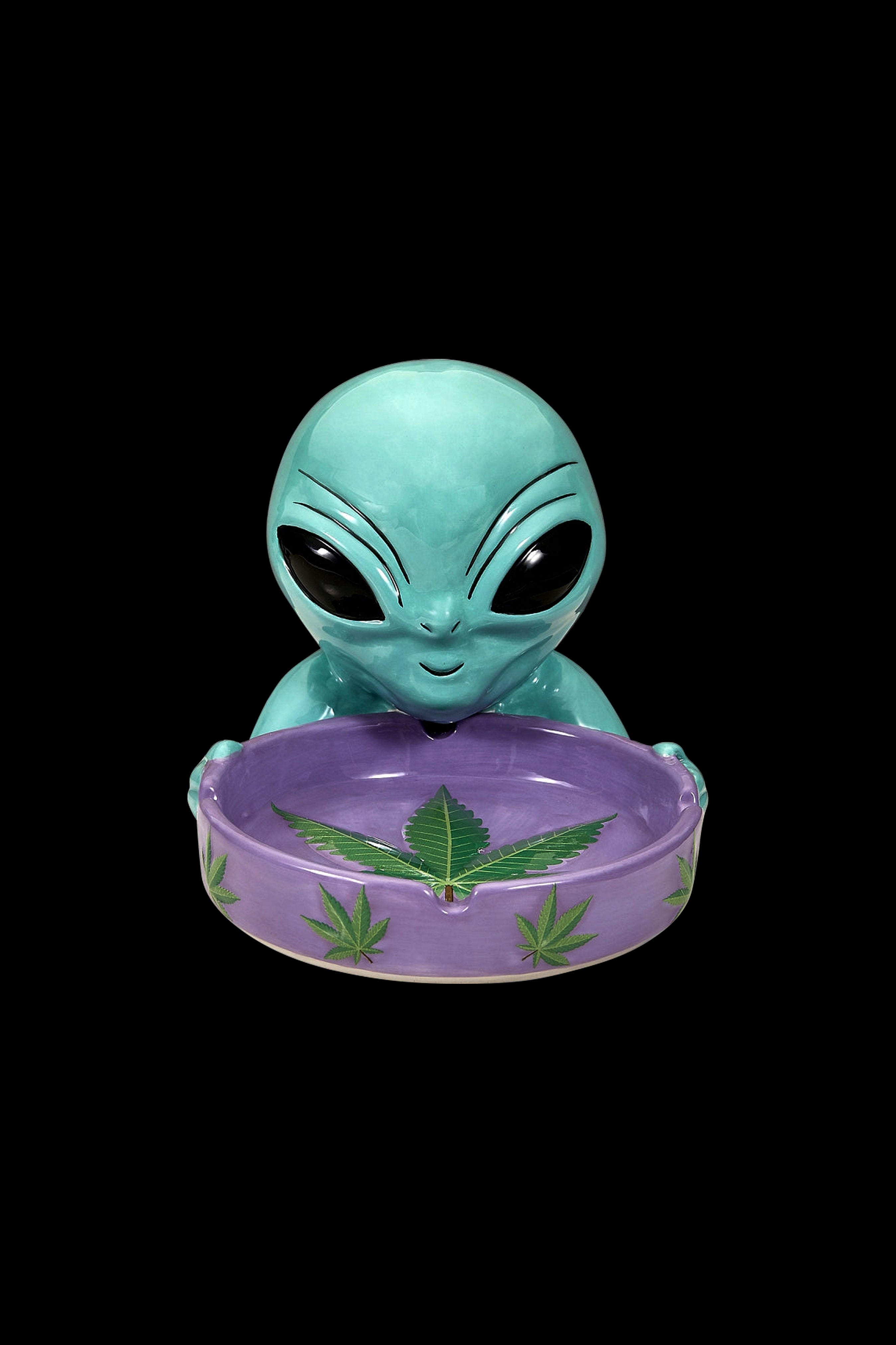 Turquoise alien head ashtray with cannabis leaf design on a purple base, isolated on black.