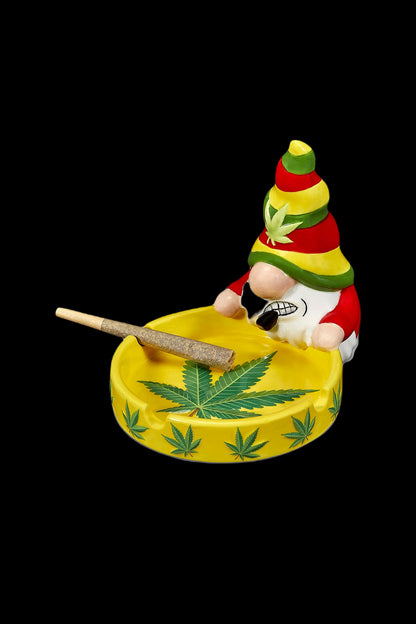 Ceramic ashtray shaped like a smiling gnome wearing a Rastafarian hat, with a lit joint and cannabis leaf inside, isolated on a black background."