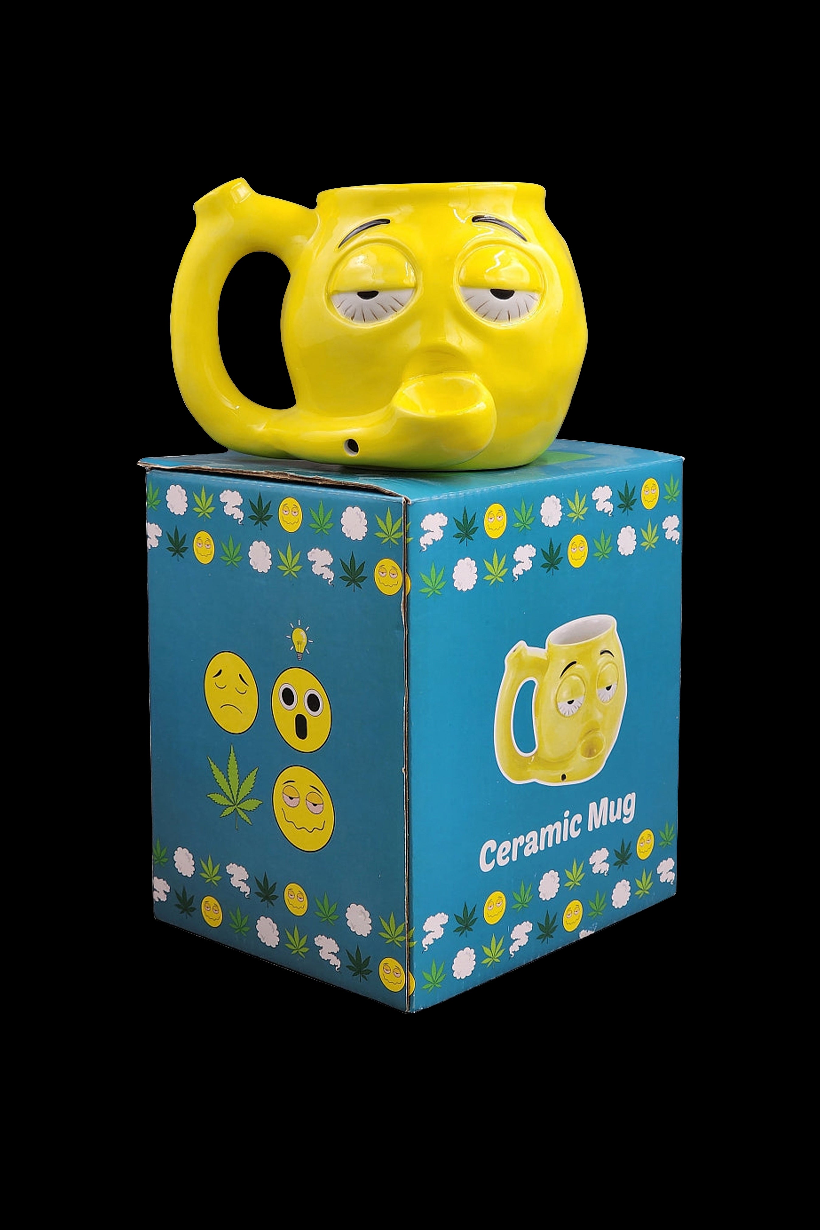 A yellow ceramic mug shaped like a relaxed face rests atop its packaging, a blue box decorated with cannabis leaves and smiling emoji faces, labeled "Ceramic Mug."