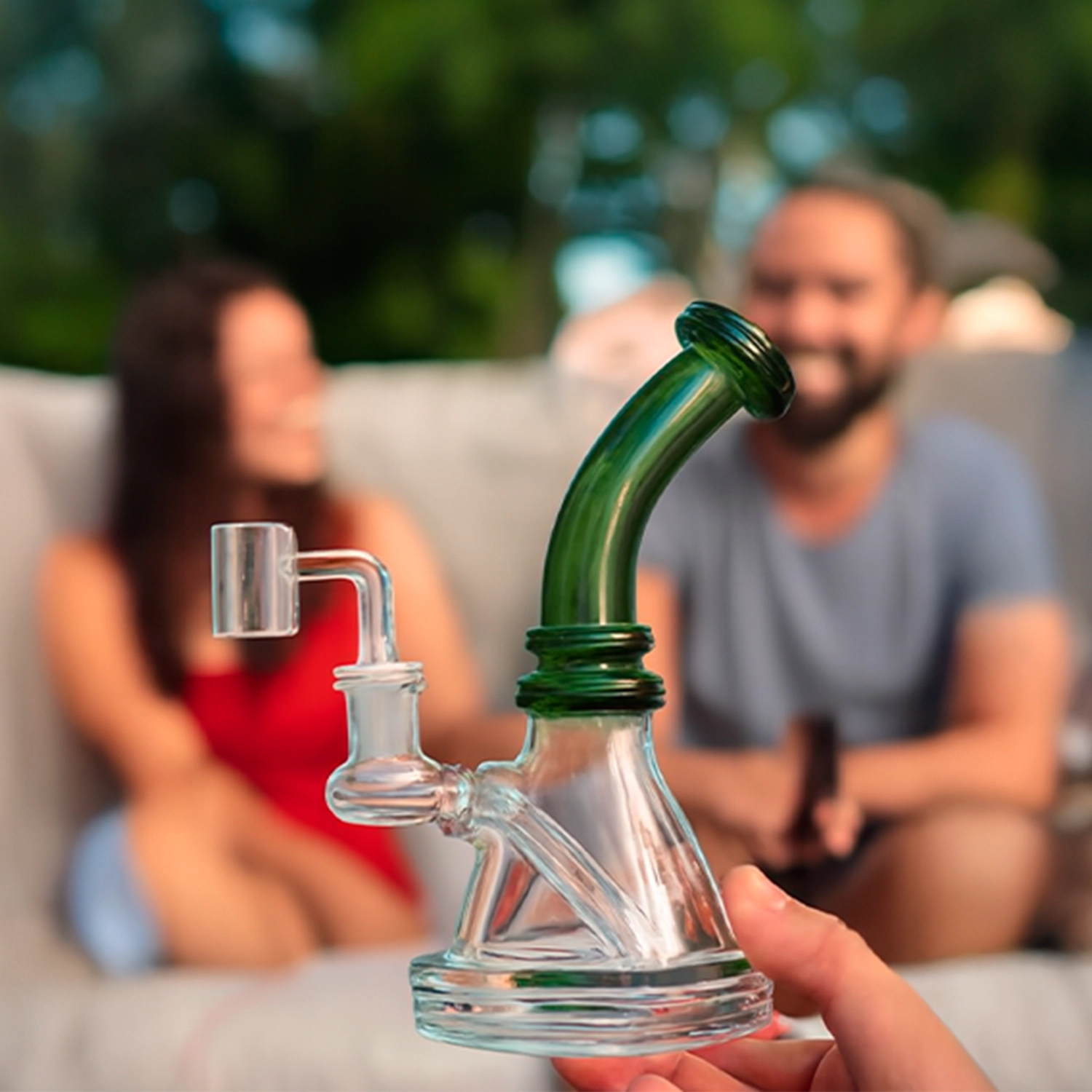 Close-up of a teal Mini Bent Neck Bong held foreground-center with its curved neck and clear base, blurred people and greenery in the background.