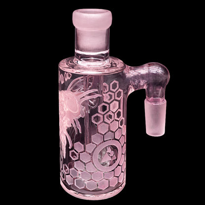 Close-up, three-quarter view of the pink Milkyway Bee Hive Wet Ash Catcher showing its frosted joint, angled side arm, and honeycomb-and-bee etching against a black background