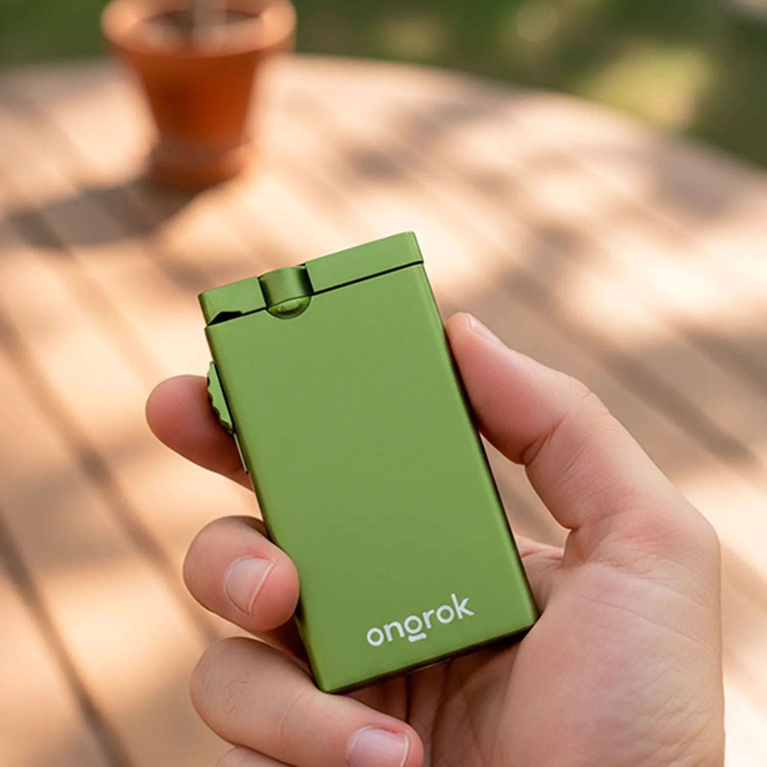 Close-up of a hand holding a matte green ONGROK Aluminum Dugout with its lid and thumb wheel visible against a sunlit deck.