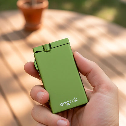 Close-up of a hand holding a matte green ONGROK Aluminum Dugout with its lid and thumb wheel visible against a sunlit deck.