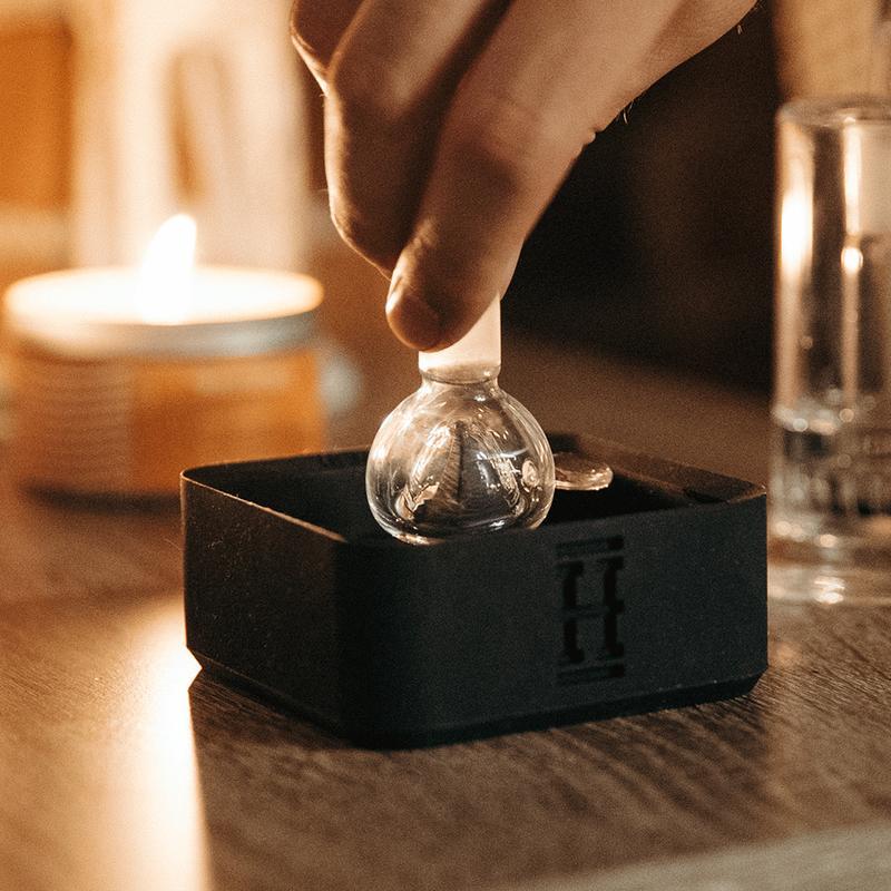 Hand placing a large ice sphere into a silicone mold, with a lit candle and glass nearby, suggesting a cannabis smoking setup.