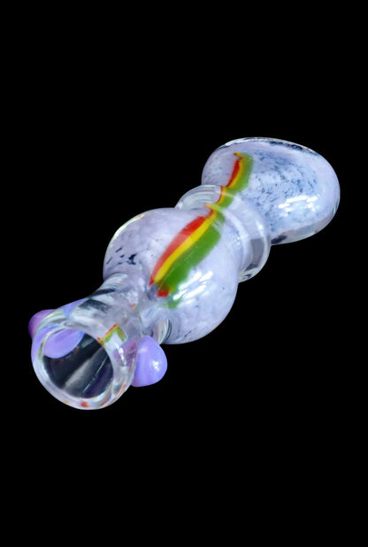 Angled close-up of the Smoke Cartel Frit Rasta One Hitter - 3 in. showing a clear glass body with purple glass knobs and a central red-yellow-green rasta stripe.