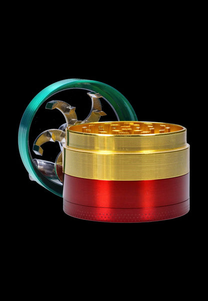 Close-up three-quarter view of the Smoke Cartel 3-Stage with Handle Grinder - 52mm showing its stacked red and gold metal chambers and a teal lid with visible silver teeth against a black background.