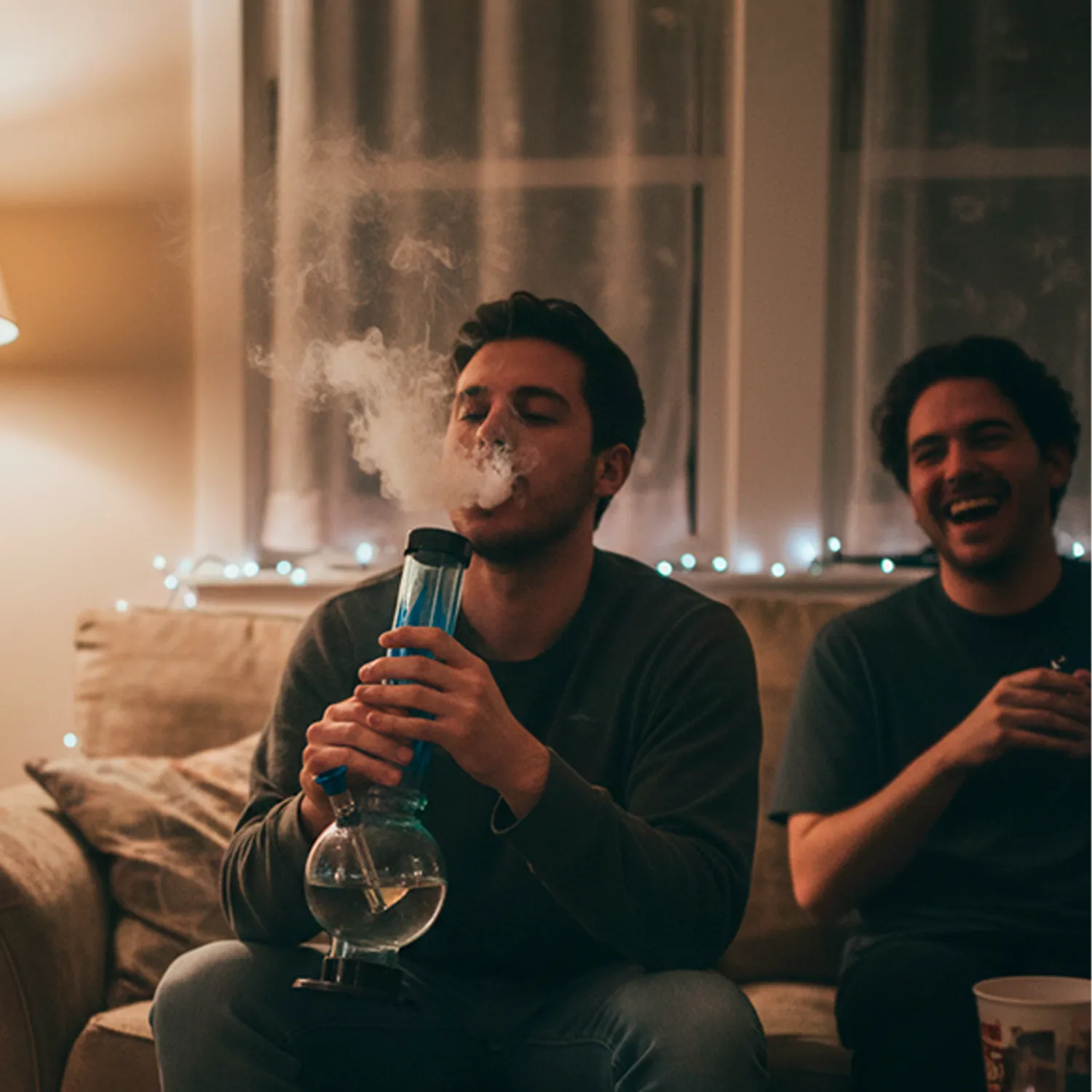 Center-framed view of a young man exhaling smoke while holding an Acrylic Bong 1.5 X 12 BUBBLE FLAME on a cozy sofa with string lights behind.