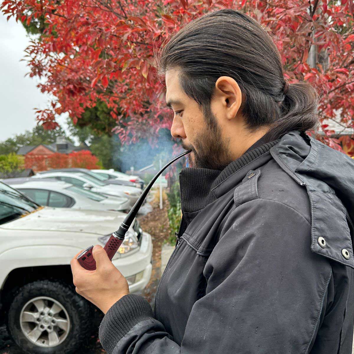 Side profile of a man outdoors using a Cipher Nautilus V3 Churchwarden Stem Kit pipe with a long curved stem and burgundy bowl against red autumn leaves and parked cars.