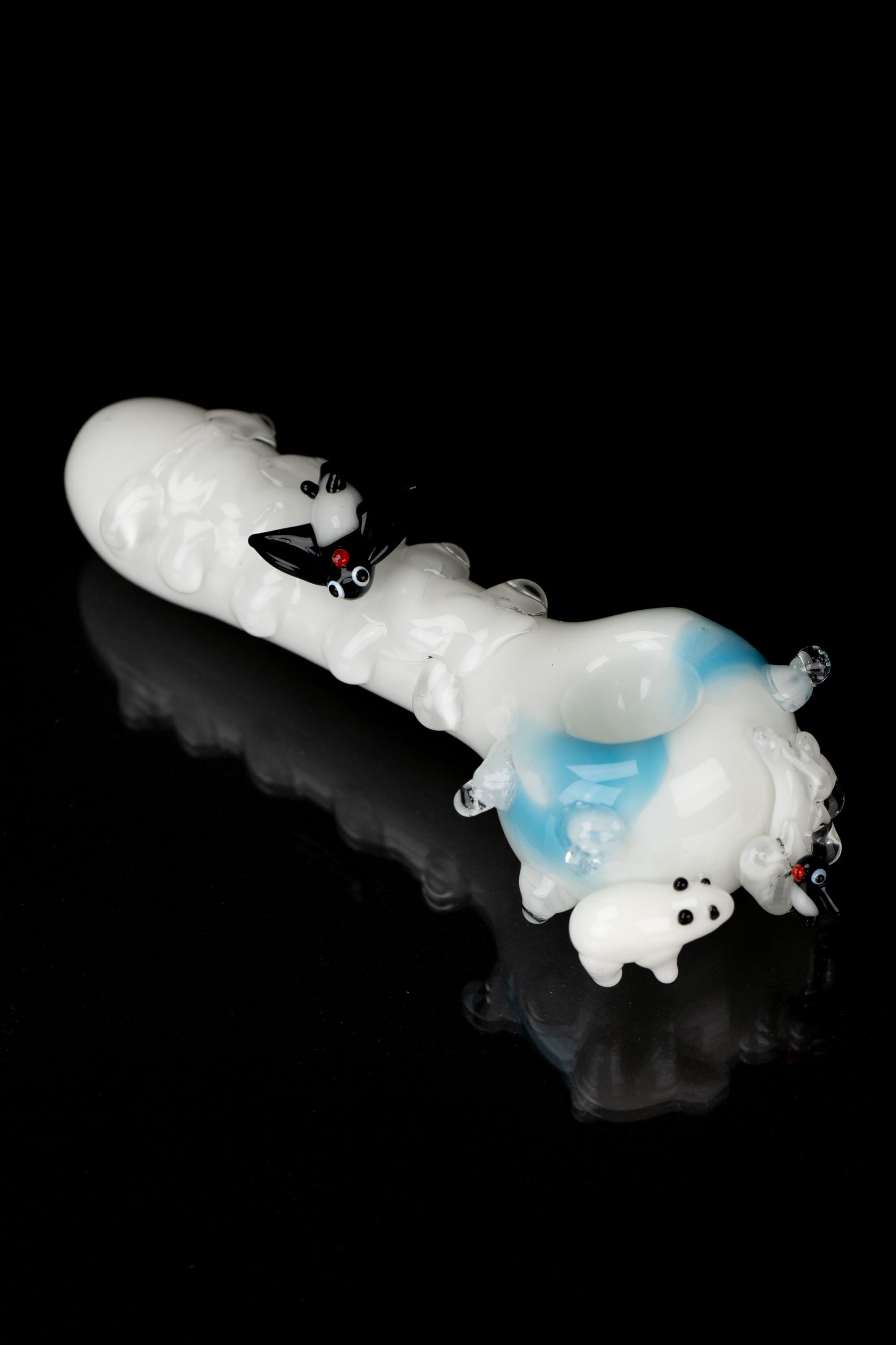 Glass pipe shaped like a white and blue cartoon dragon with red eyes, isolated on a black reflective surface.