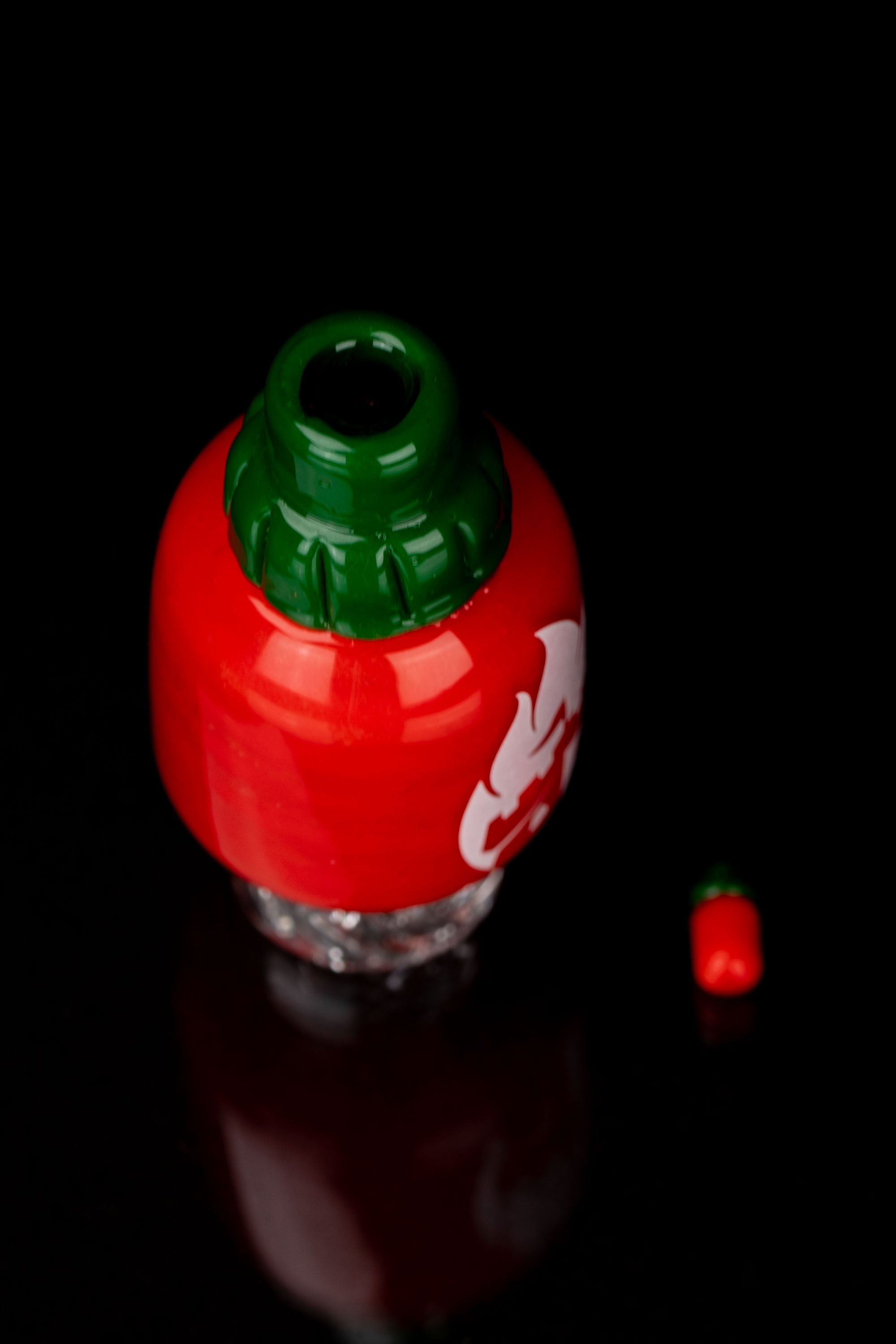 Red strawberry-shaped glass piece with a green leafy top, likely a cannabis water pipe, against a black background.