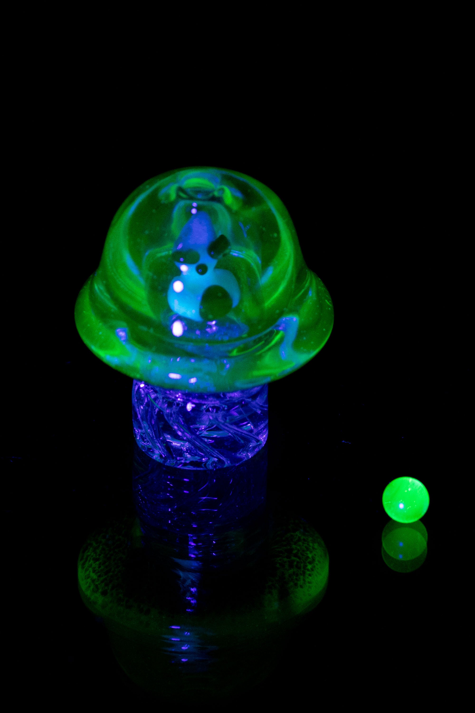 A vibrant, glowing green and blue glass mushroom-shaped dab rig with accompanying glass marbles, illuminated against a black background.