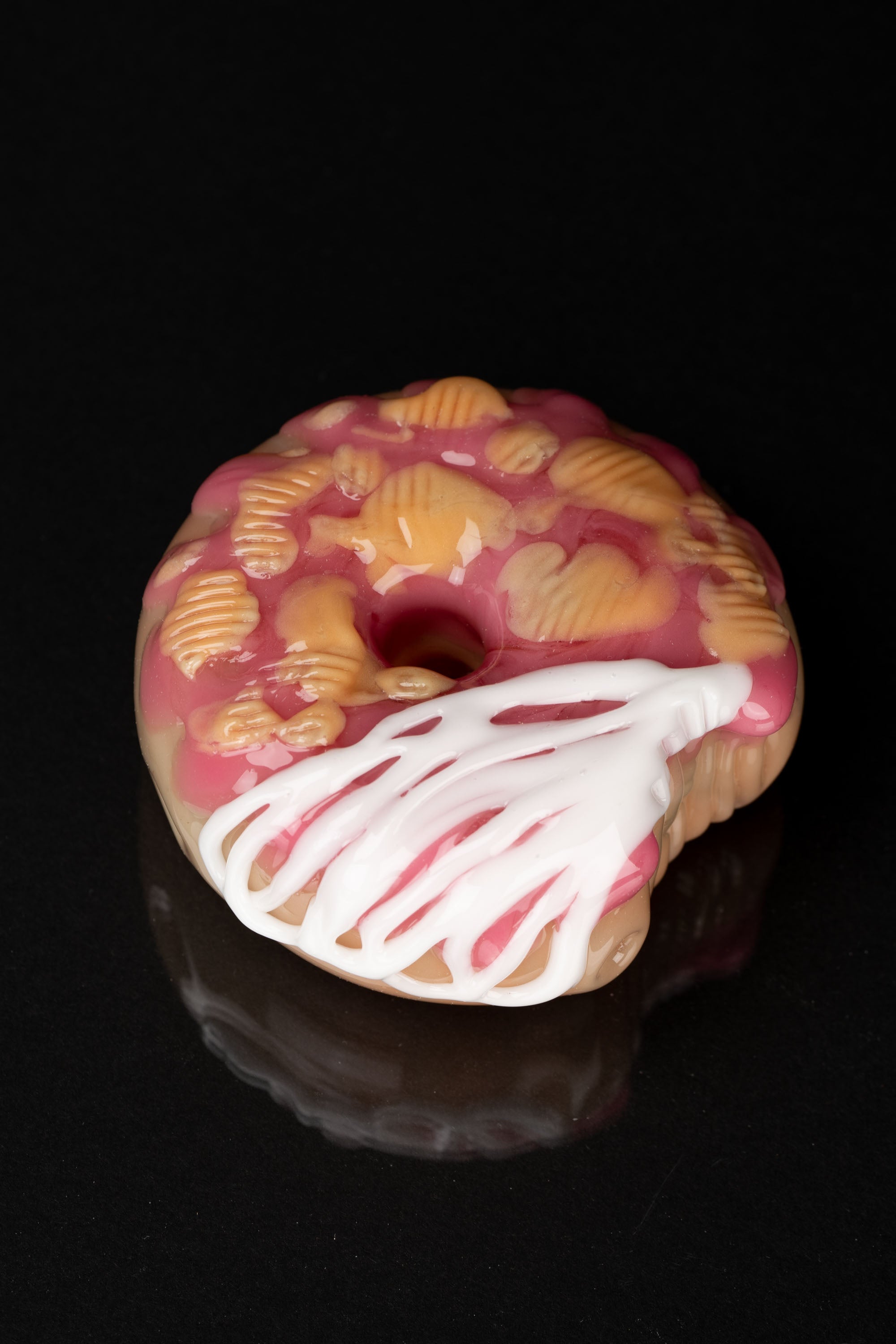 Pink glass donut-shaped smoking piece with detailed frosting and pastry designs, emitting smoke against a black background.