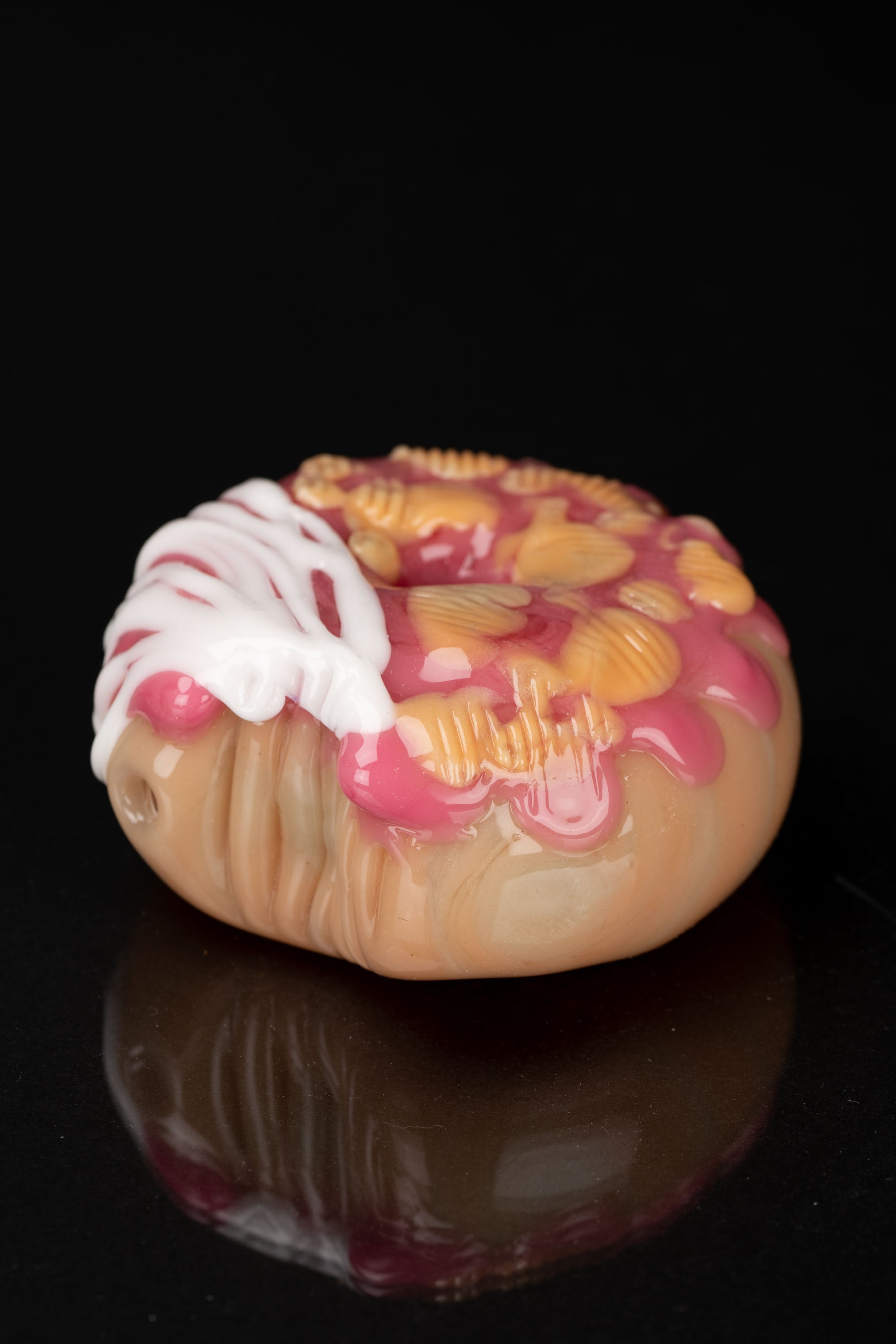 Hand-blown glass cannabis pipe shaped like a glazed donut with strawberry topping, reflecting on a dark surface.