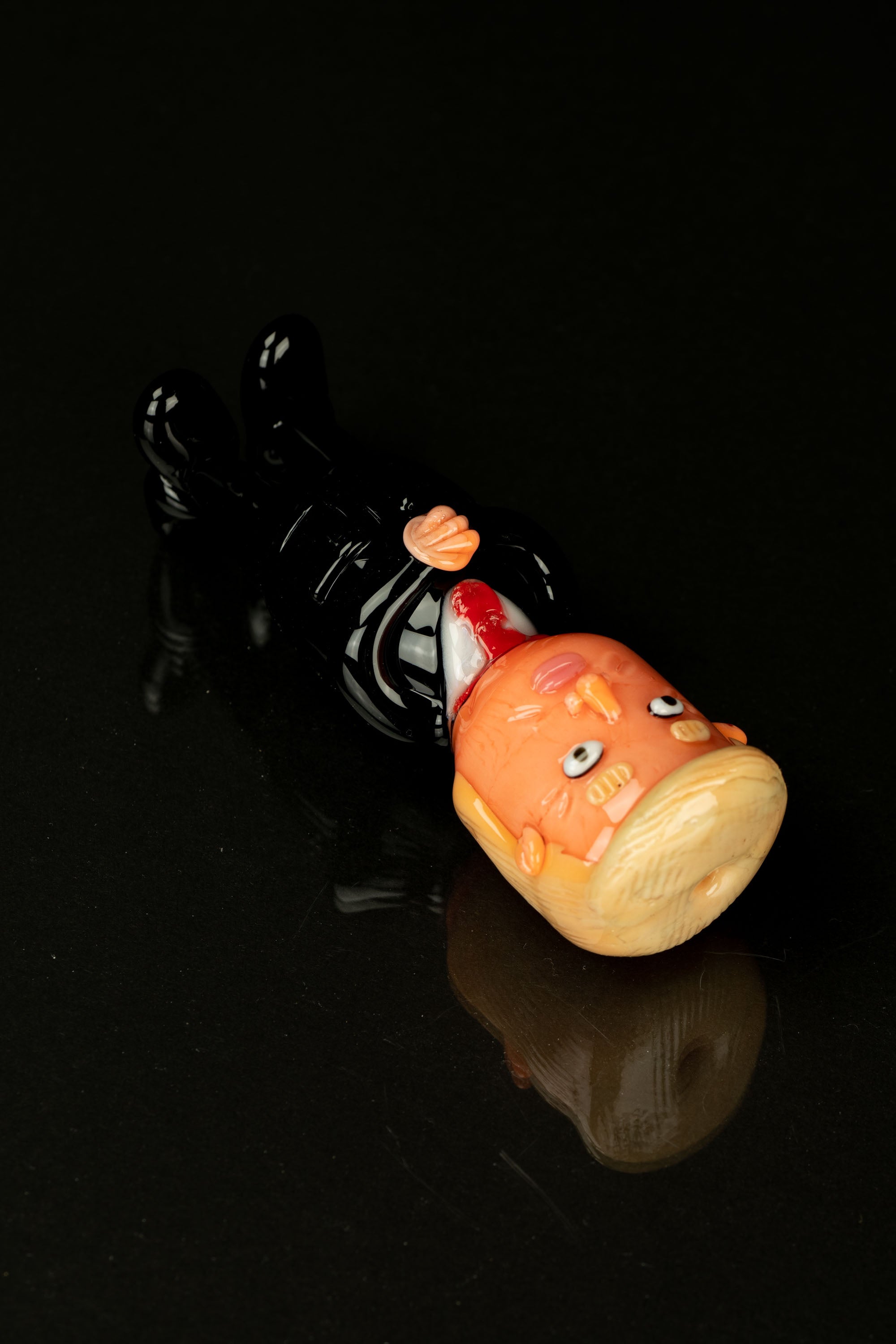 Glass sculpted cannabis bud with a comical, cartoonish face, resting on a black glass surface with a reflective sheen.