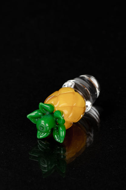 Glass cannabis pipe shaped like a pineapple with green leaves, displayed on a reflective surface against a black background.
