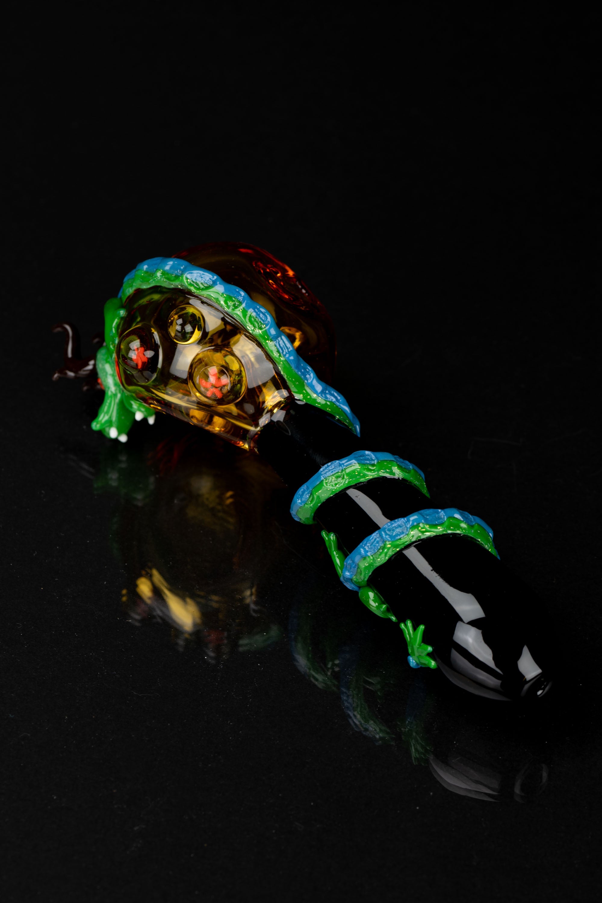 Intricately designed glass cannabis pipe with a vibrant green and blue pattern, displayed against a black reflective surface.