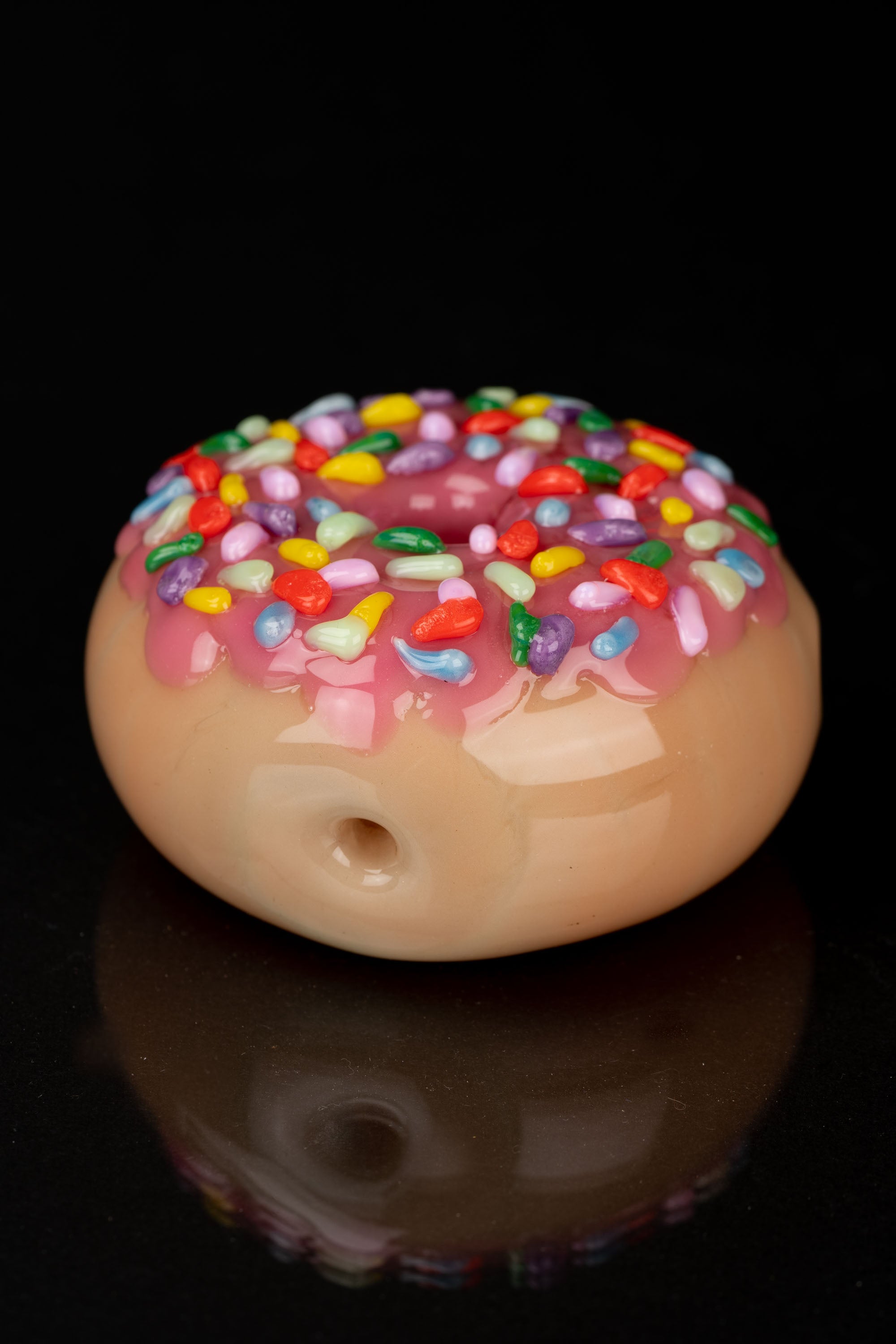 Glass cannabis water pipe shaped like a frosted donut with colorful sprinkles, reflecting on a dark surface.