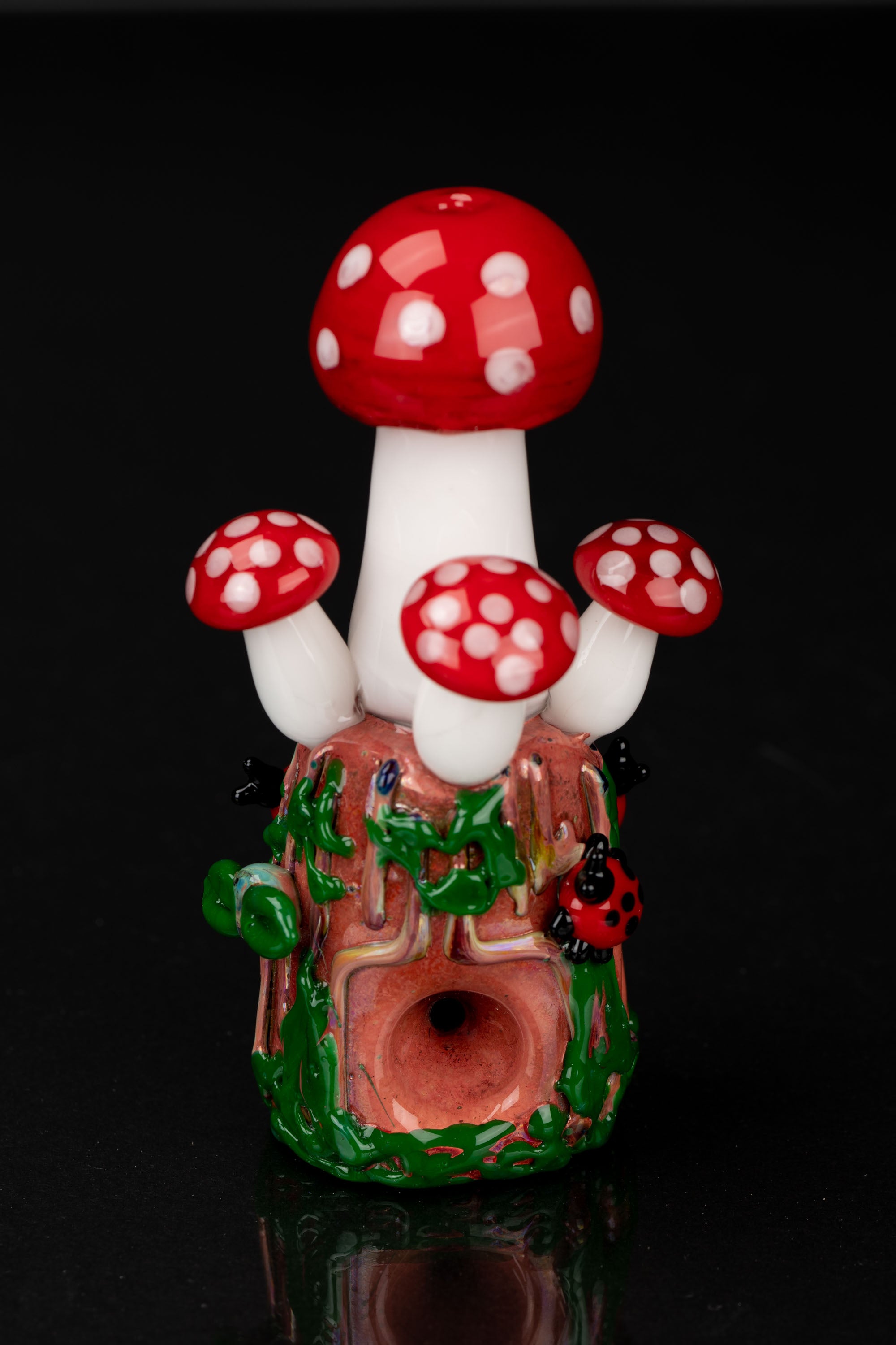 Hand-blown glass mushroom-shaped cannabis pipe with red and white spotted caps and green foliage detail, reflecting on a dark surface.