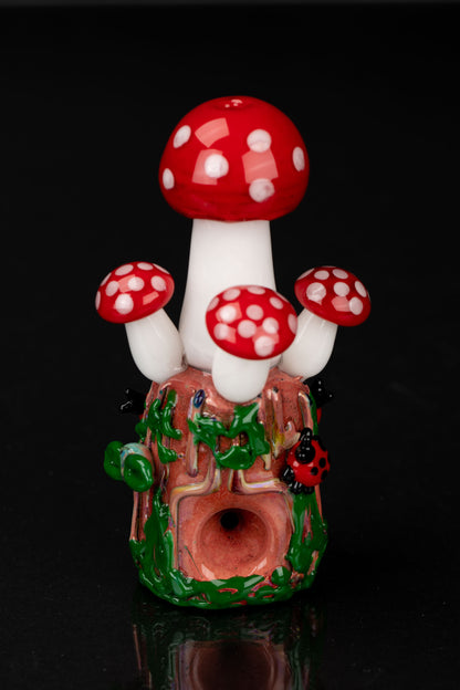 Hand-blown glass mushroom-shaped cannabis pipe with red and white spotted caps and green foliage detail, reflecting on a dark surface.
