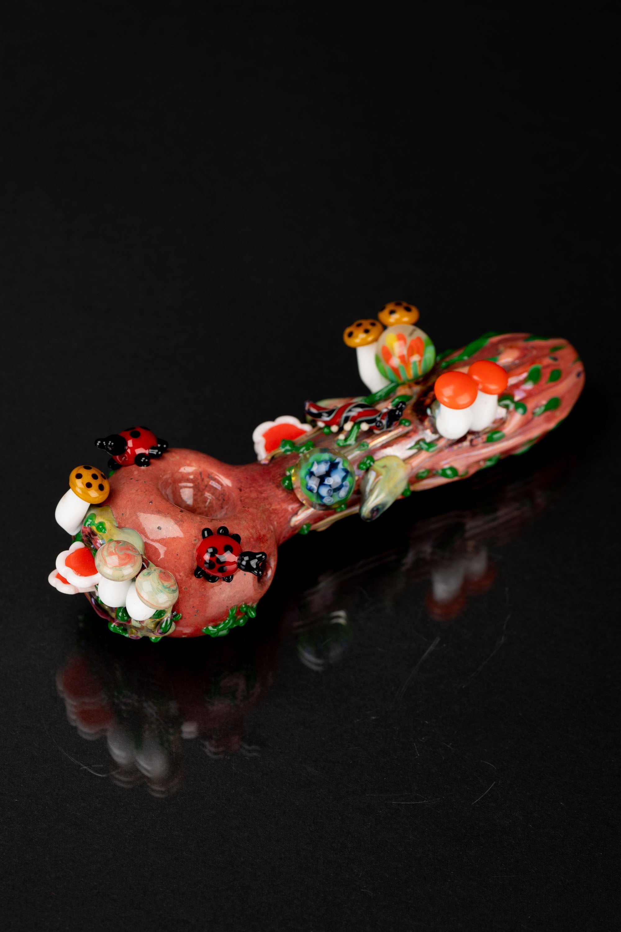 Hand-blown glass pipe decorated with colorful mushroom designs, reflecting on a dark surface.