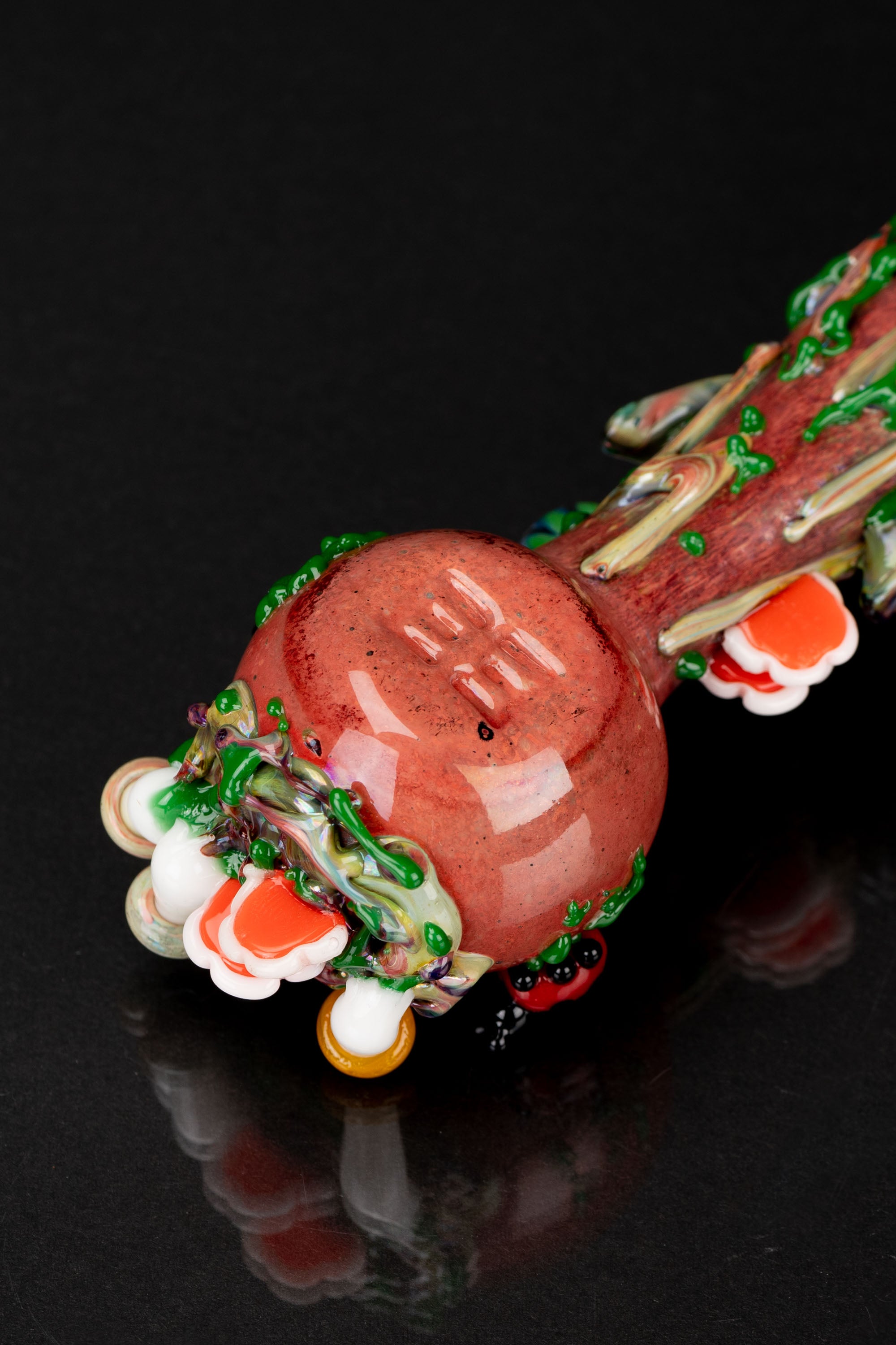 Close-up of a colorful, hand-blown glass cannabis pipe with a bulbous, reddish-orange chamber and intricate green and white details, reflecting on a dark surface.