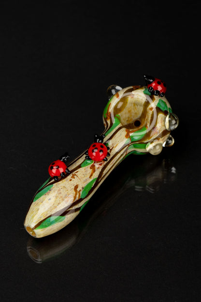 Angled close-up of the Empire Glassworks Lady Bug Mini Spoon Pipe showing its speckled tan and green glass body with three small red ladybug accents on a black background