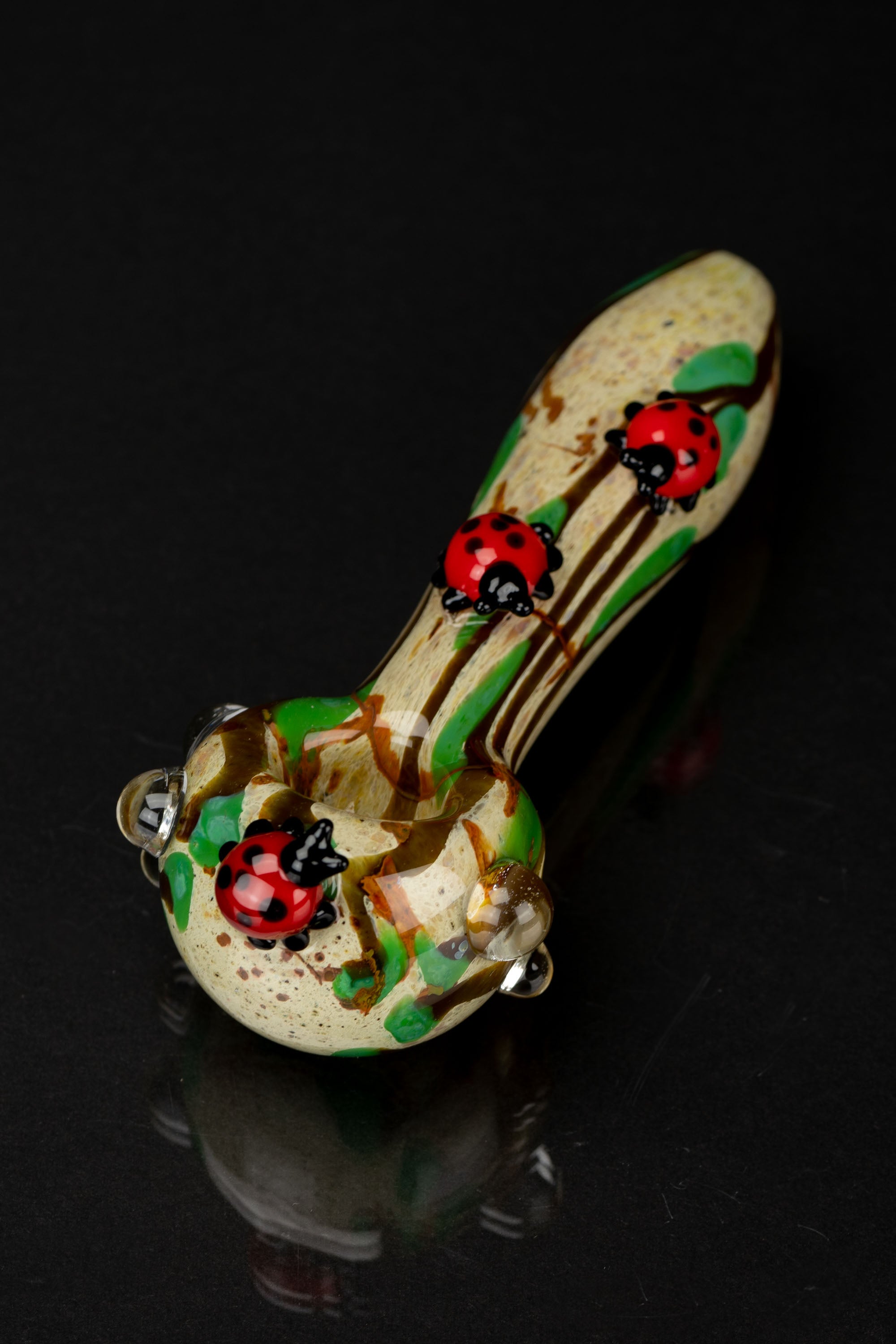 Angled close-up of the Empire Glassworks Lady Bug Mini Spoon Pipe showing two glossy red ladybug decorations on a speckled beige and green glass body against a black background