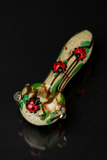 Angled close-up of the Empire Glassworks Lady Bug Mini Spoon Pipe showing two glossy red ladybug decorations on a speckled beige and green glass body against a black background