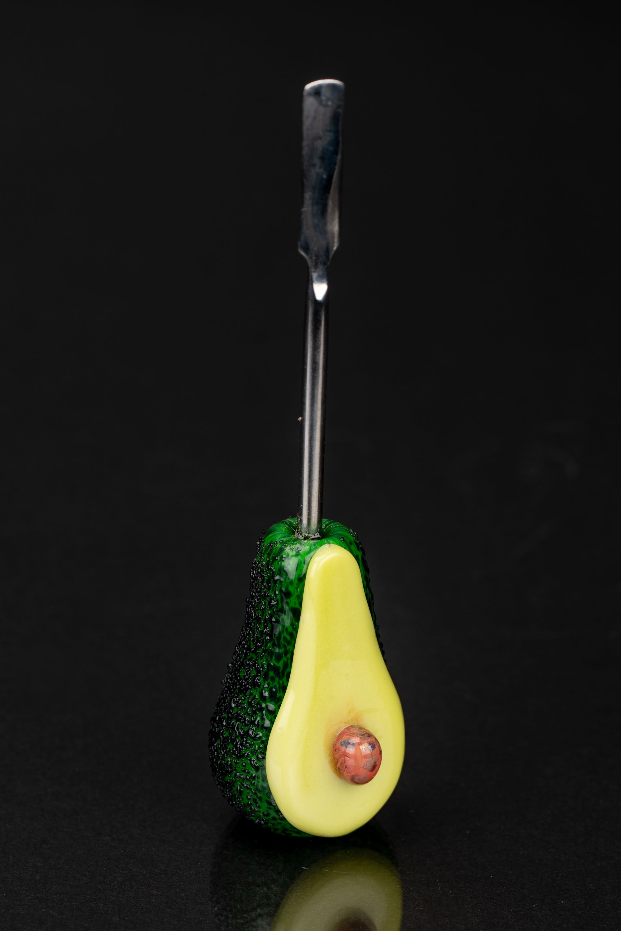 Glass avocado-shaped dab tool with a metallic scoop, reflecting on a dark surface.