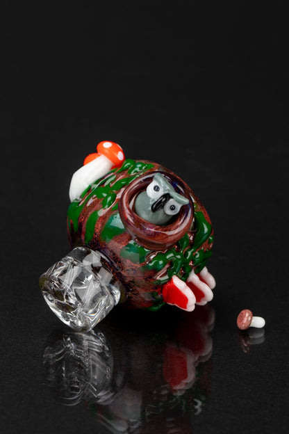 A glass pipe shaped like a cartoon frog with red and green swirls, resting on a reflective black surface alongside a small glass bead.