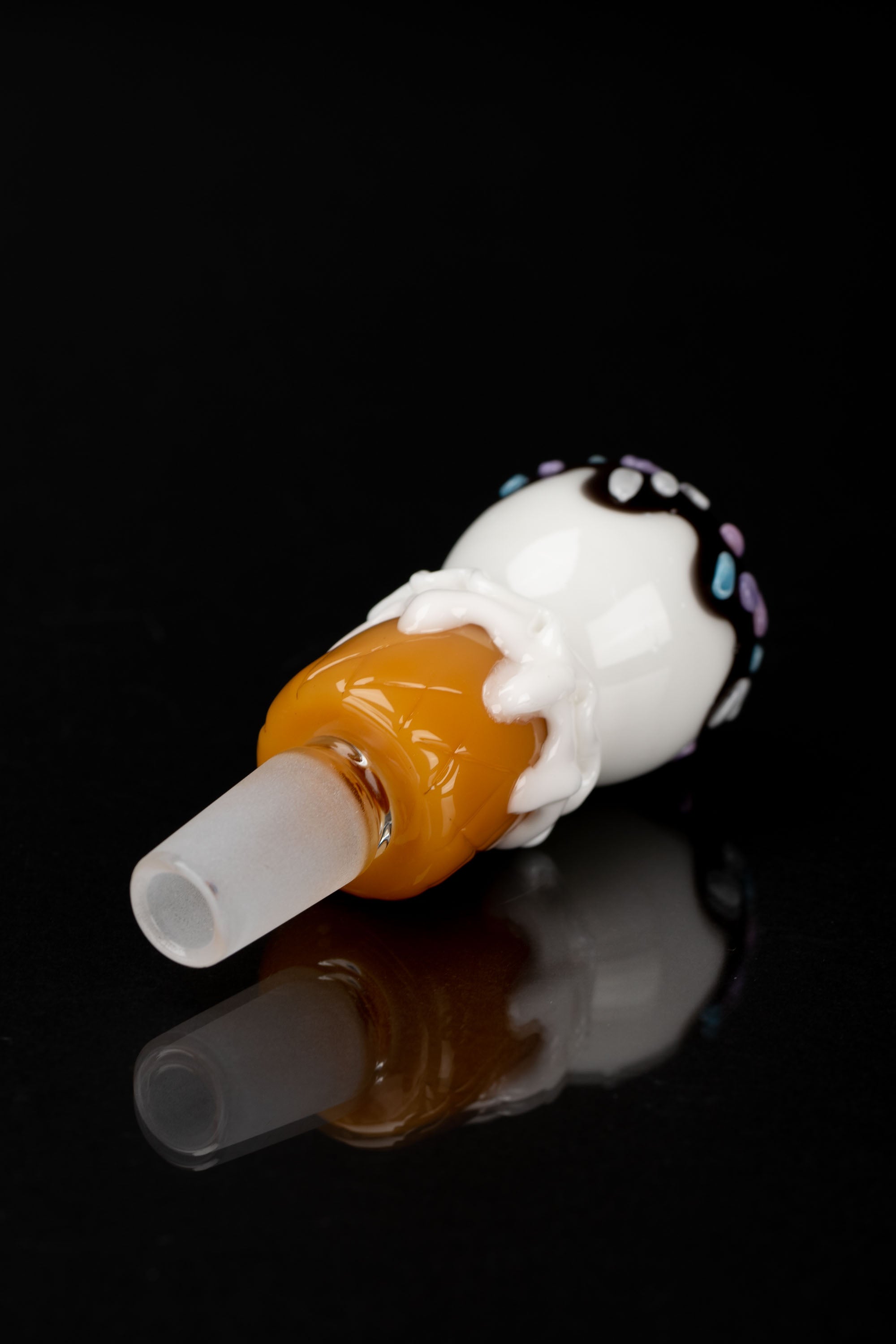 A glass cannabis smoking piece shaped like a melting ice cream cone with colorful sprinkles, reflecting on a black surface.